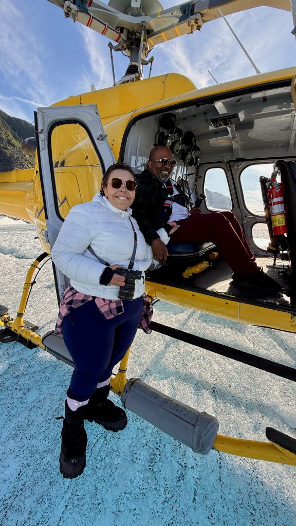 A man and woman pose with a yellow helicopter, on top of a glacier. The man is seated in the helicopter and the woman is standing outside the vehicle.